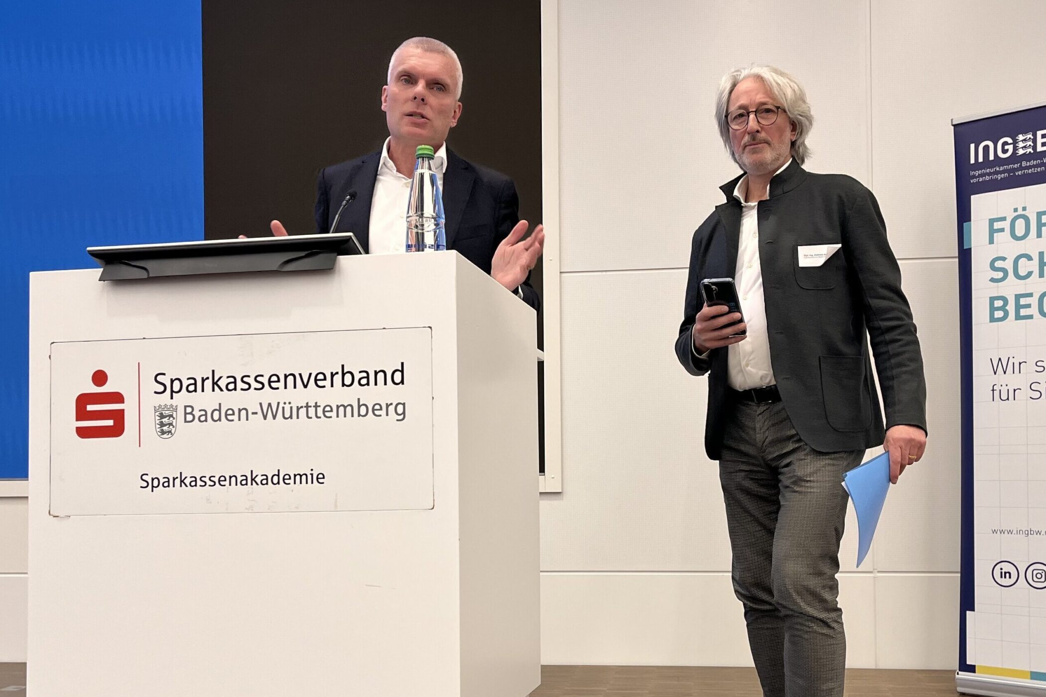 Zwei Männer bei einer Präsentation, einer am Rednerpult, der andere mit Handy.