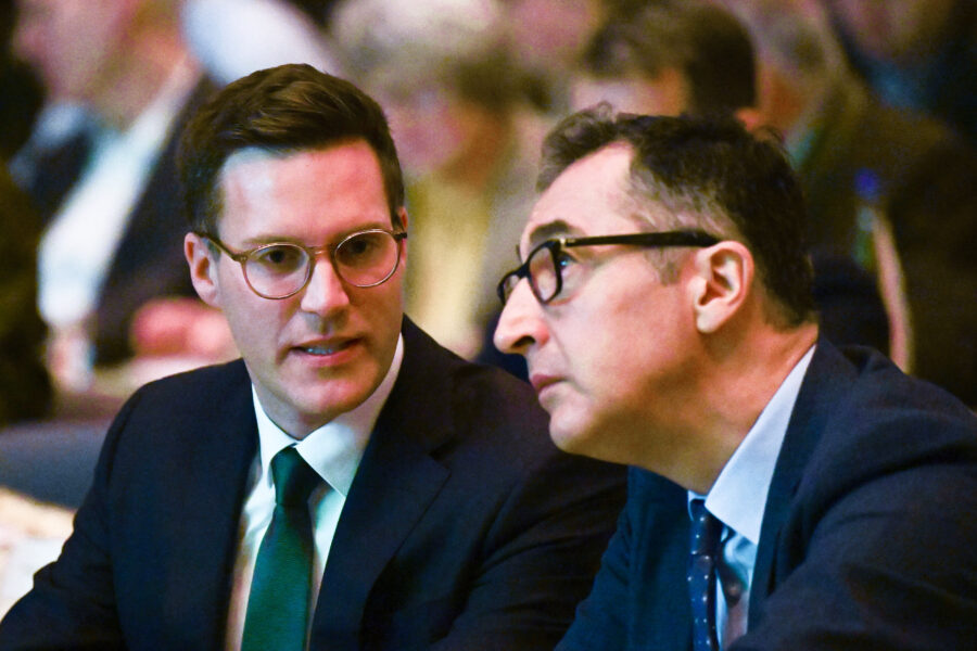 Zwei Männer in Anzügen unterhalten sich in einem vollen Raum.