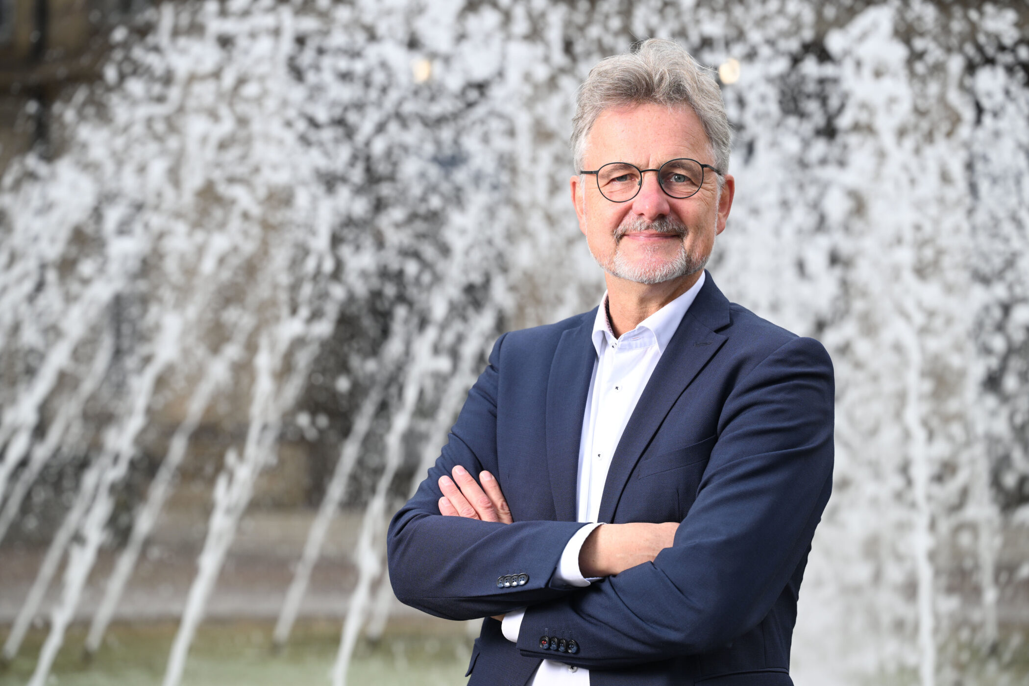 Älterer Mann mit Brille und Anzug steht vor einem sprudelnden Brunnen.