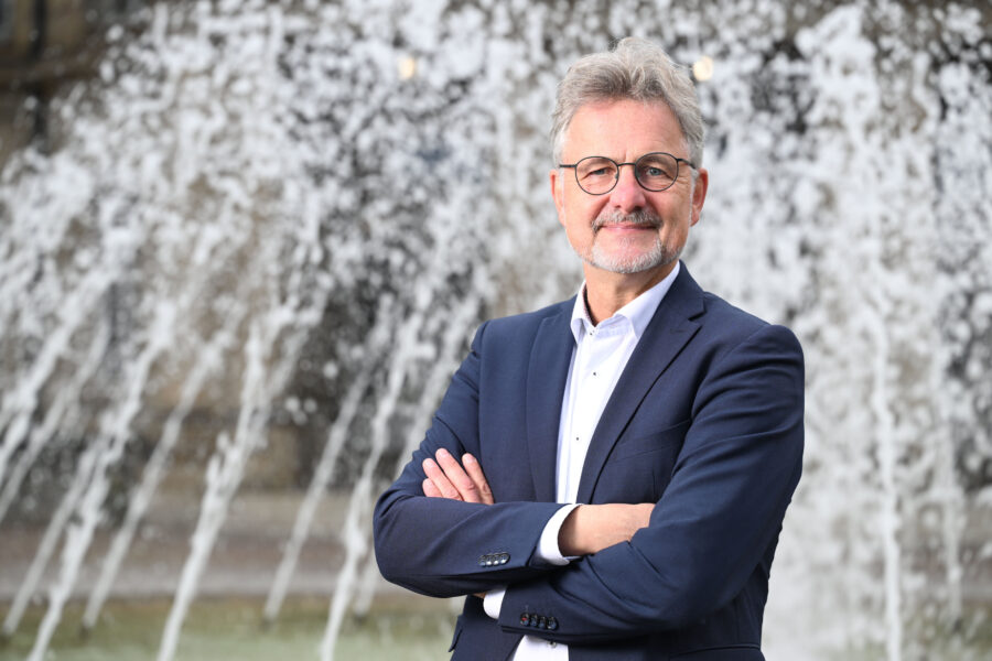 Älterer Mann mit Brille und Anzug steht vor einem sprudelnden Brunnen.