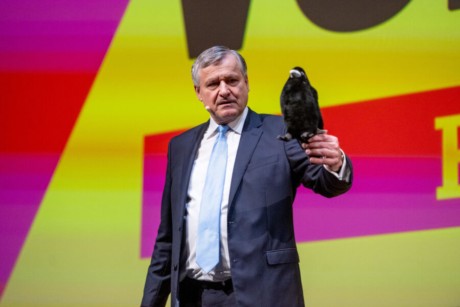 Älterer Mann im Anzug hält einen schwarzen Vogel vor buntem Hintergrund.