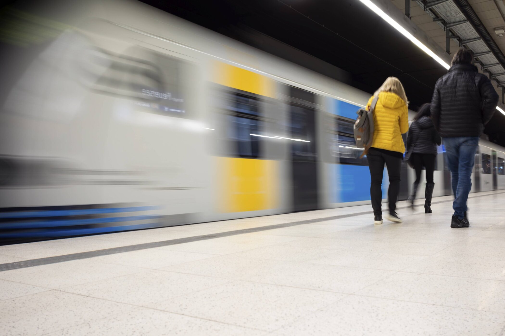 Zwei Personen gehen auf einem Bahnsteig, vorbeifahrender Zug im Hintergrund.