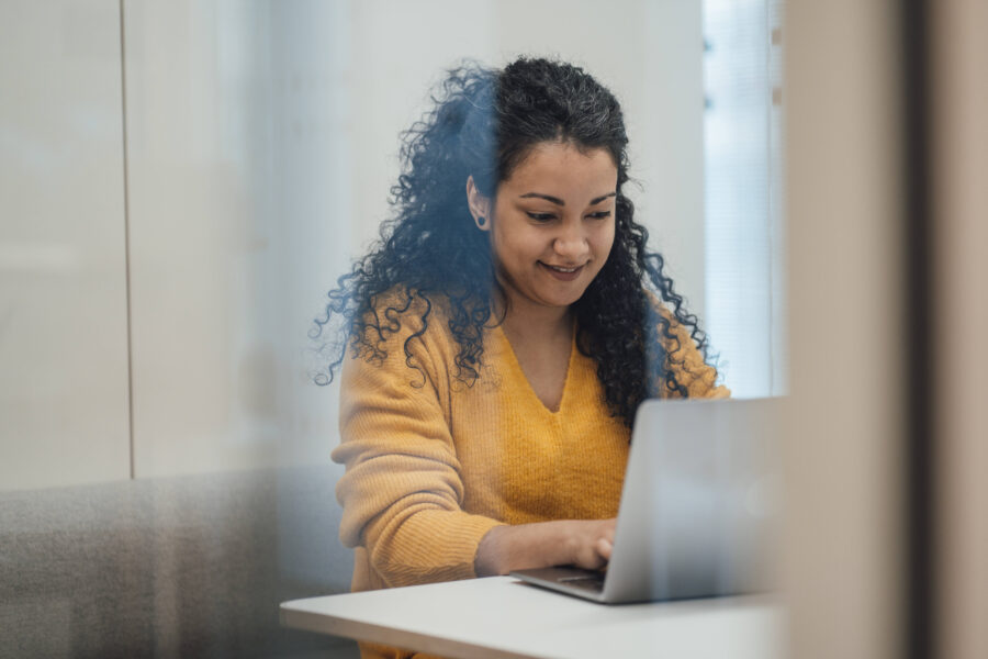 Frau in gelbem Pullover arbeitet an einem Laptop in einem Büro.