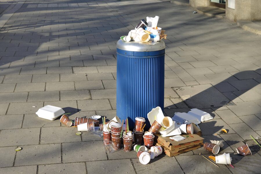 Überfüllter Mülleimer mit Müll auf dem Boden, darunter Becher und Pizzakartons.