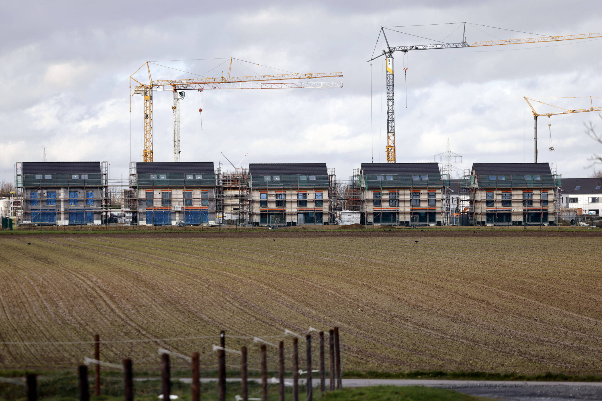 Fünf im Bau befindliche Häuser mit Kränen im Hintergrund, davor ein Feld.