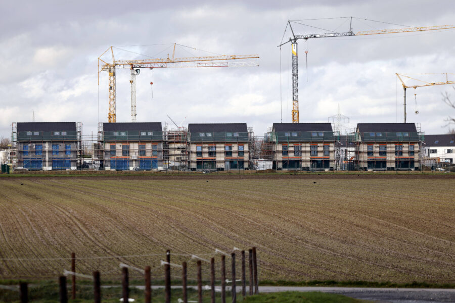 Fünf im Bau befindliche Häuser mit Kränen im Hintergrund, davor ein Feld.