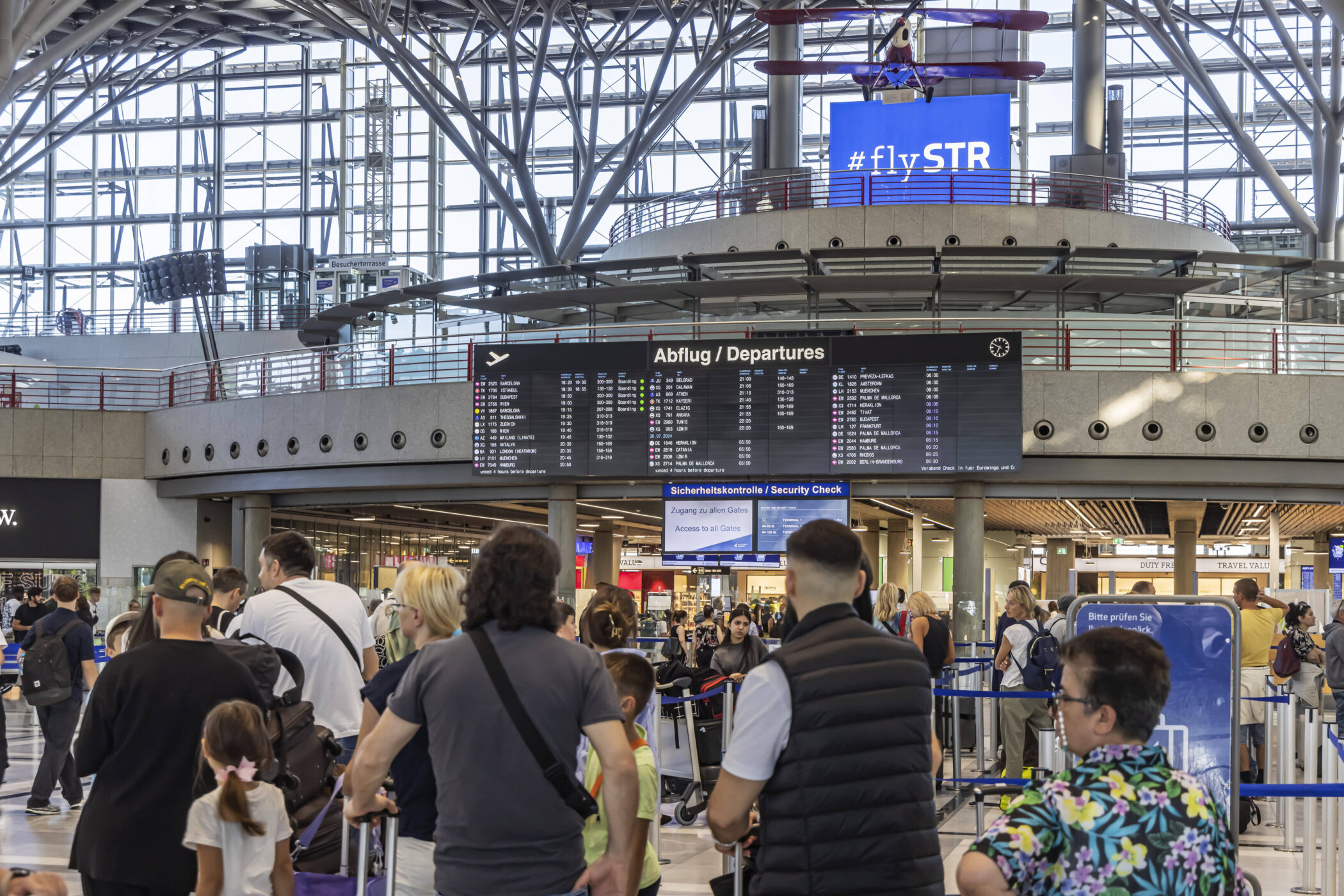 Menschenmenge am Flughafen, Abflugtafel und Sicherheitskontrolle sichtbar.