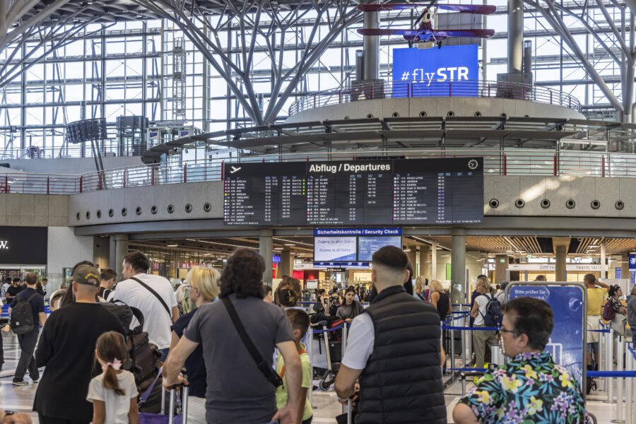 Menschenmenge am Flughafen, Abflugtafel und Sicherheitskontrolle sichtbar.