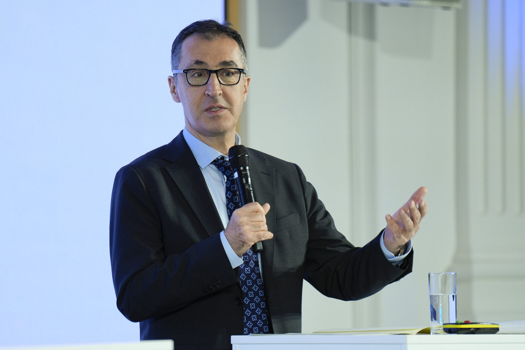 Mann im Anzug spricht in Mikrofon, Glas Wasser und Stift auf Tisch.