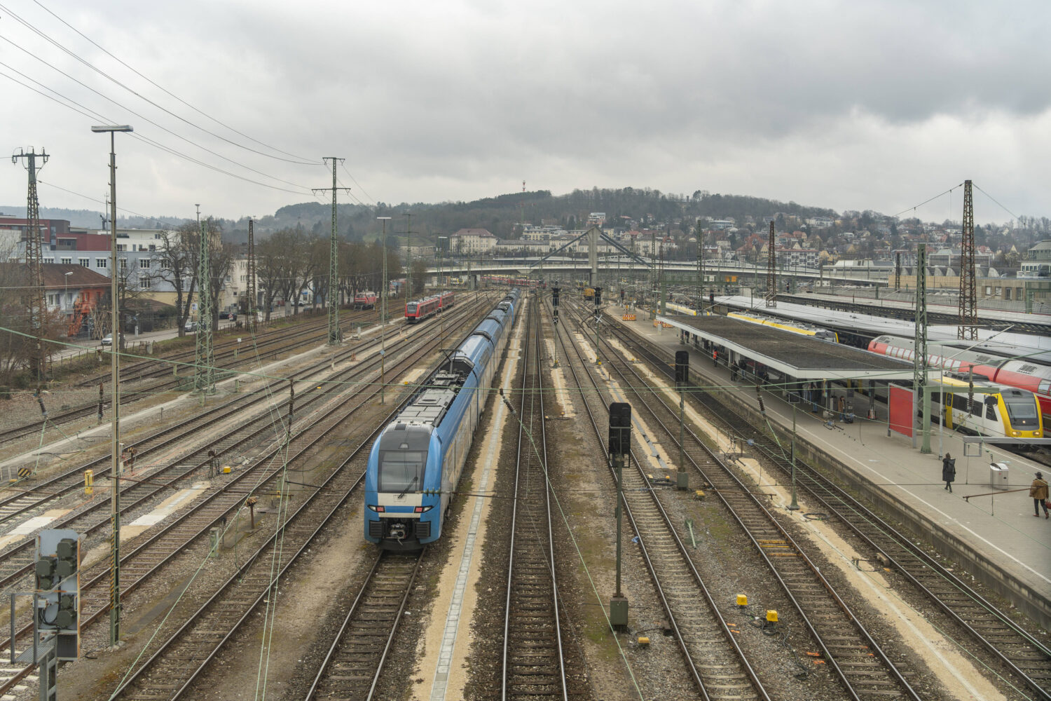 Bahnhof mit mehreren Gleisen, ein blauer Zug fährt, Menschen auf dem Bahnsteig.