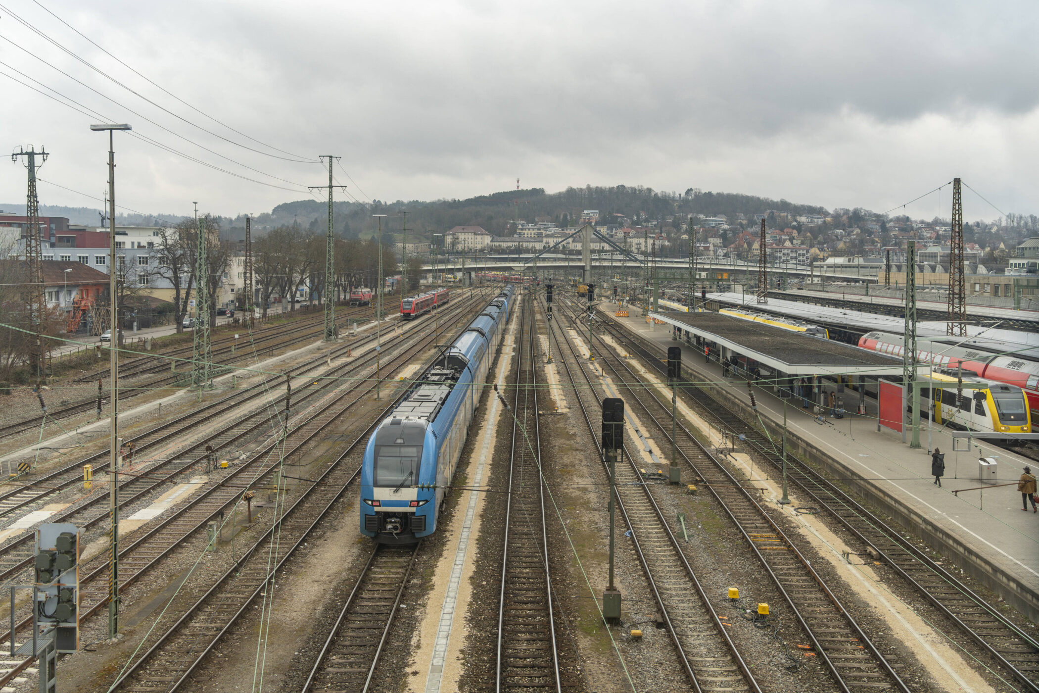 Bahnhof mit mehreren Gleisen, ein blauer Zug fährt, Menschen auf dem Bahnsteig.