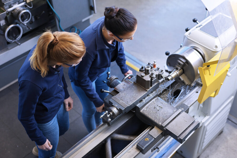 Zwei Personen arbeiten an einer Drehmaschine in einer Werkstatt.