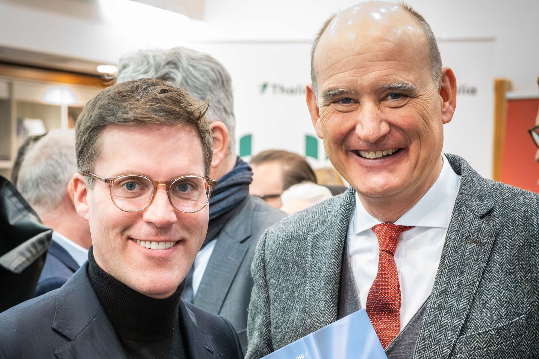 Zwei lächelnde Männer in Anzügen, einer hält ein Buch.