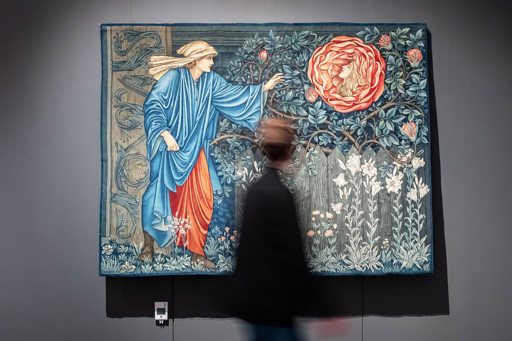 Person geht an einem Wandteppich mit Blumenmotiv vorbei.