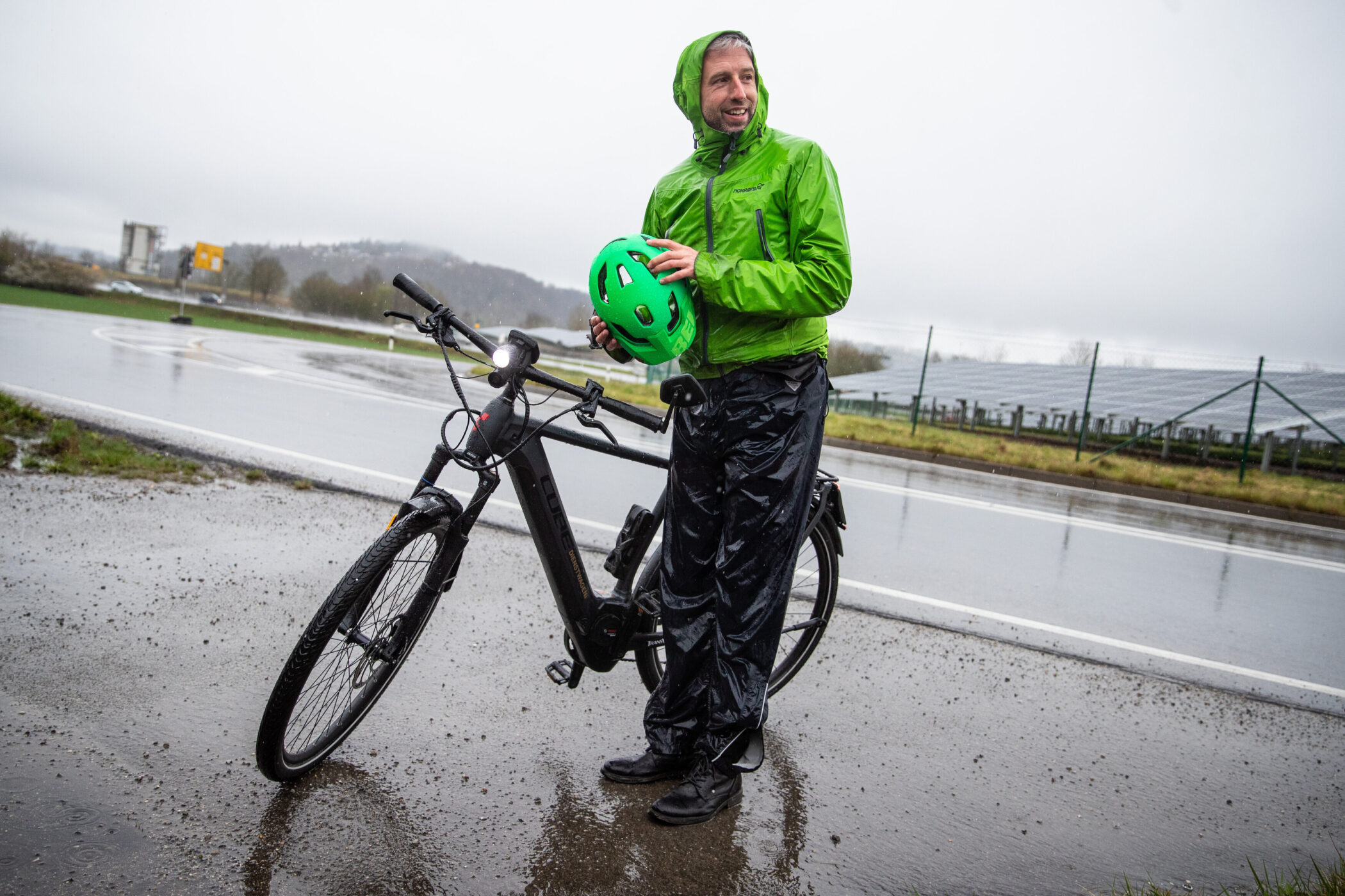 Mann in grüner Regenjacke hält Helm neben Fahrrad auf nasser Straße.