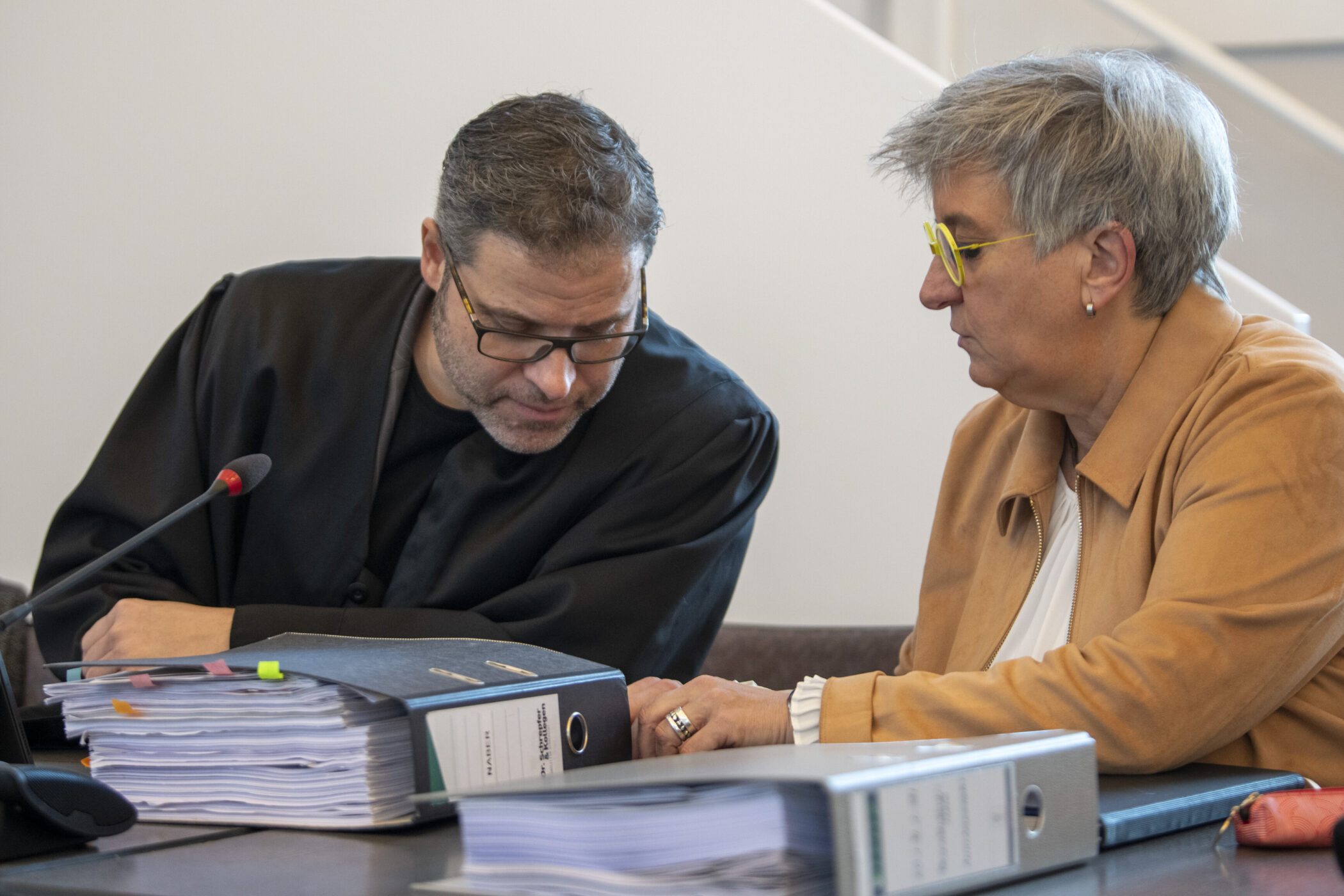 Zwei Personen sitzen an einem Tisch mit Aktenordnern und sprechen miteinander.