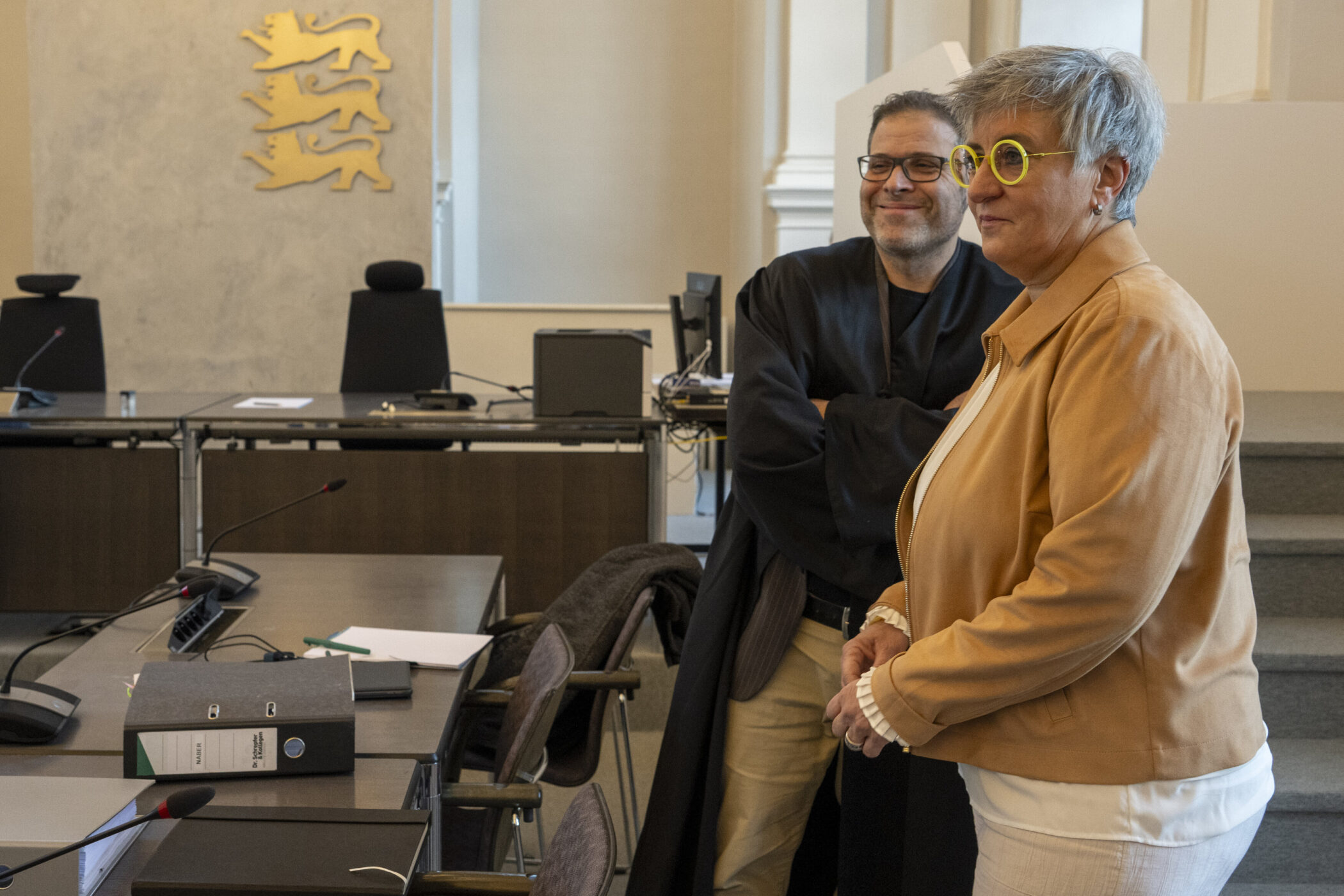 Zwei Personen stehen in einem Raum mit Tischen und Stühlen, eine trägt eine Robe.