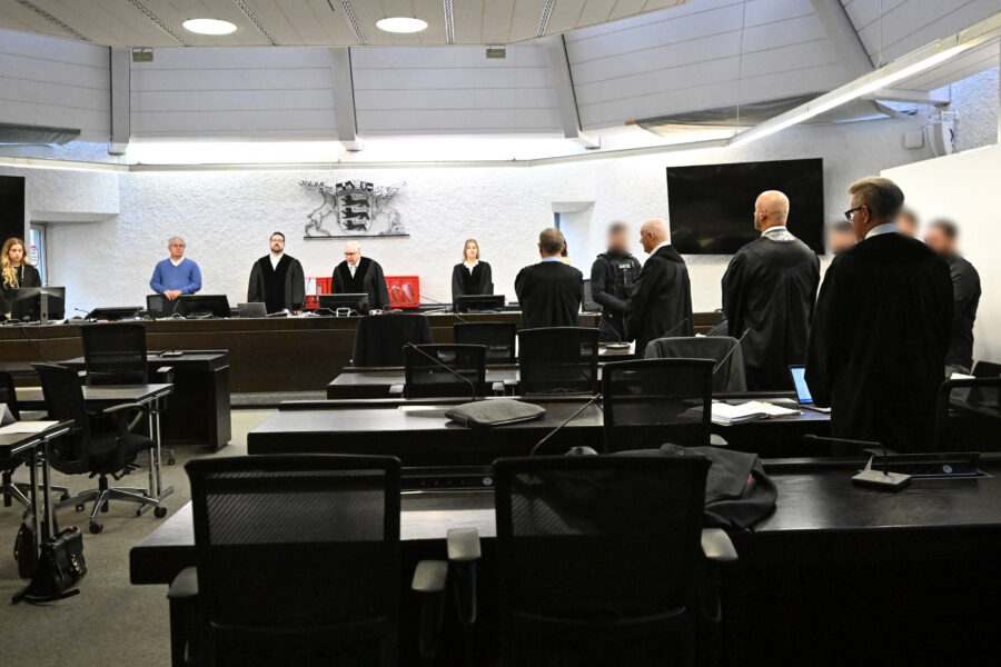 Gerichtssaal mit stehenden Personen in Roben, leere Stühle im Vordergrund.