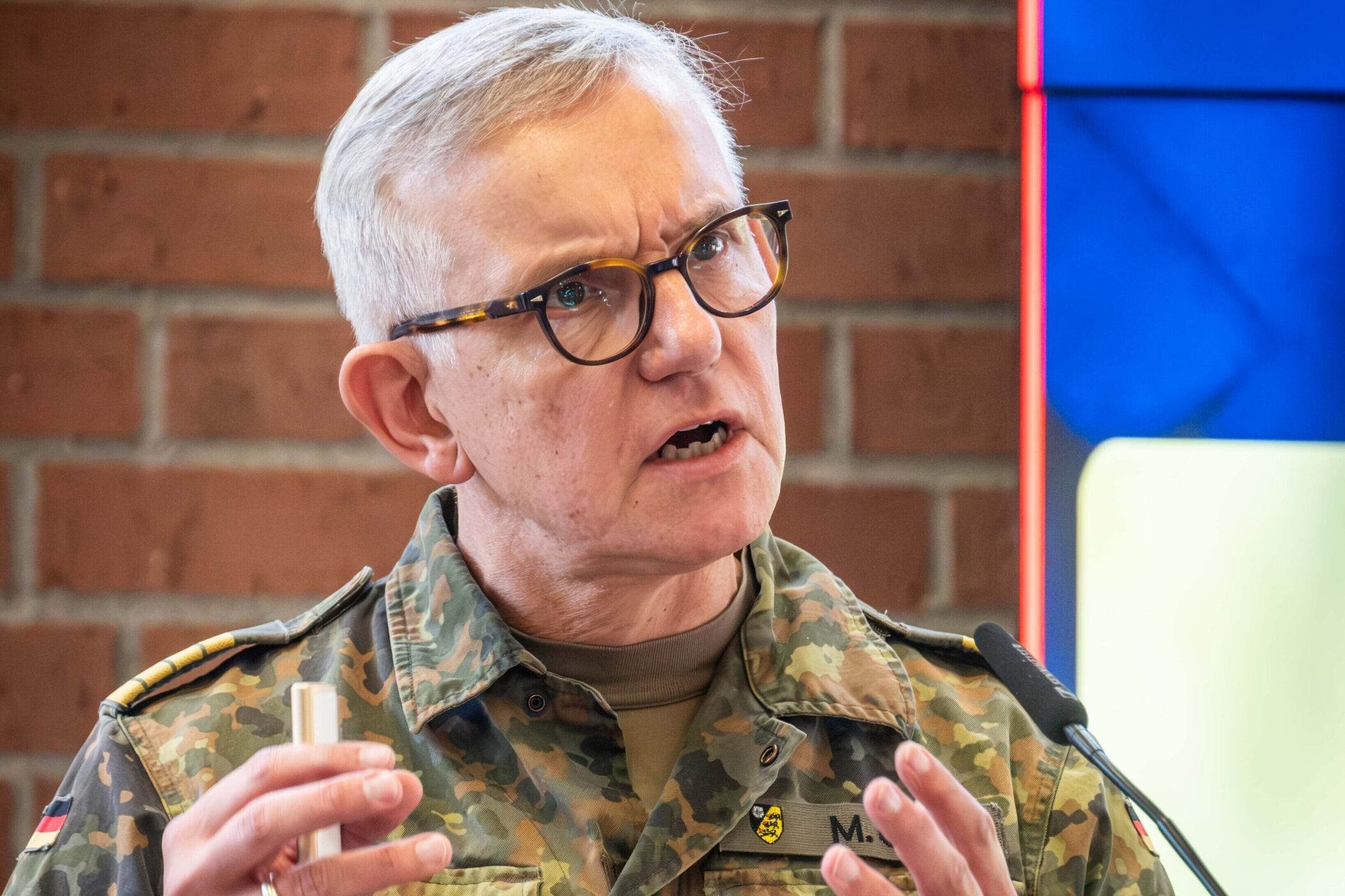 Älterer Mann in Uniform spricht in ein Mikrofon, Backsteinwand im Hintergrund.