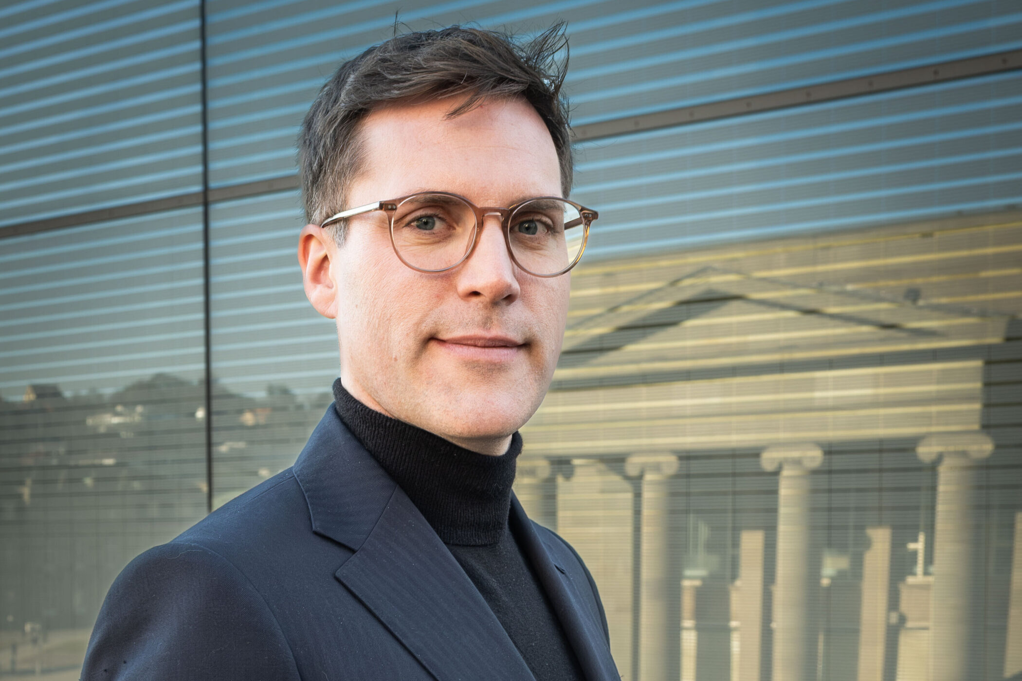 Mann mit Brille und Anzug vor Glasfassade mit Säulenreflexion.