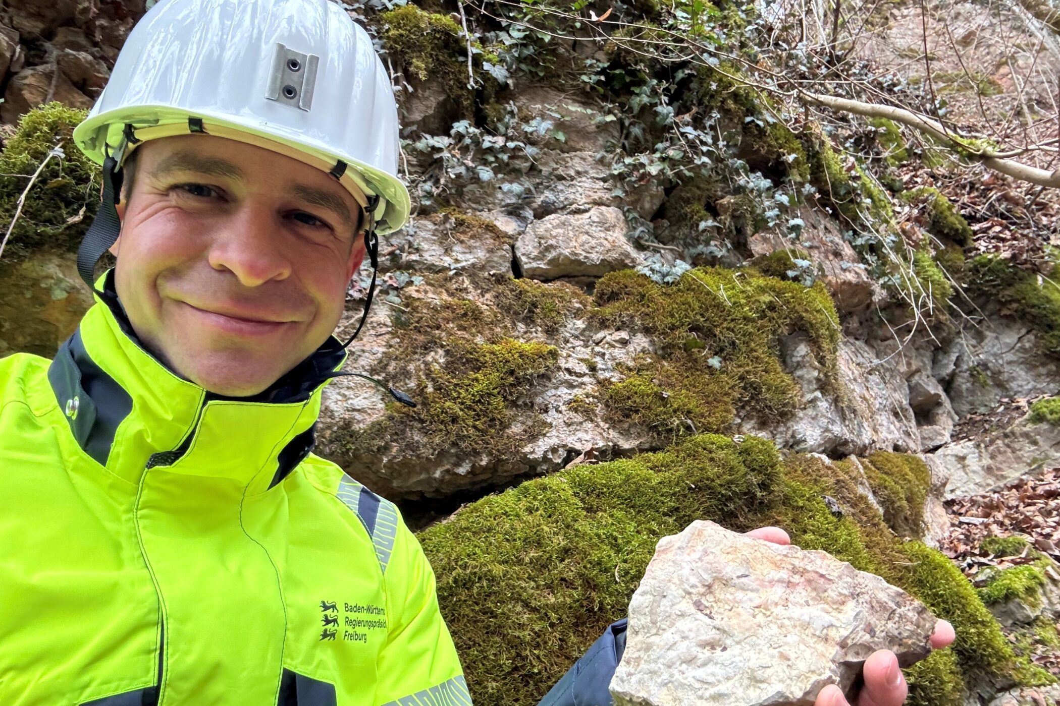 Person mit Helm und gelber Jacke hält einen Stein vor moosbewachsener Felswand.