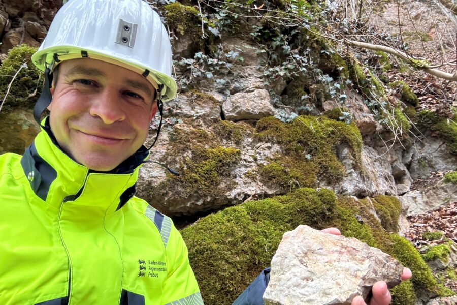 Person mit Helm und gelber Jacke hält einen Stein vor moosbewachsener Felswand.