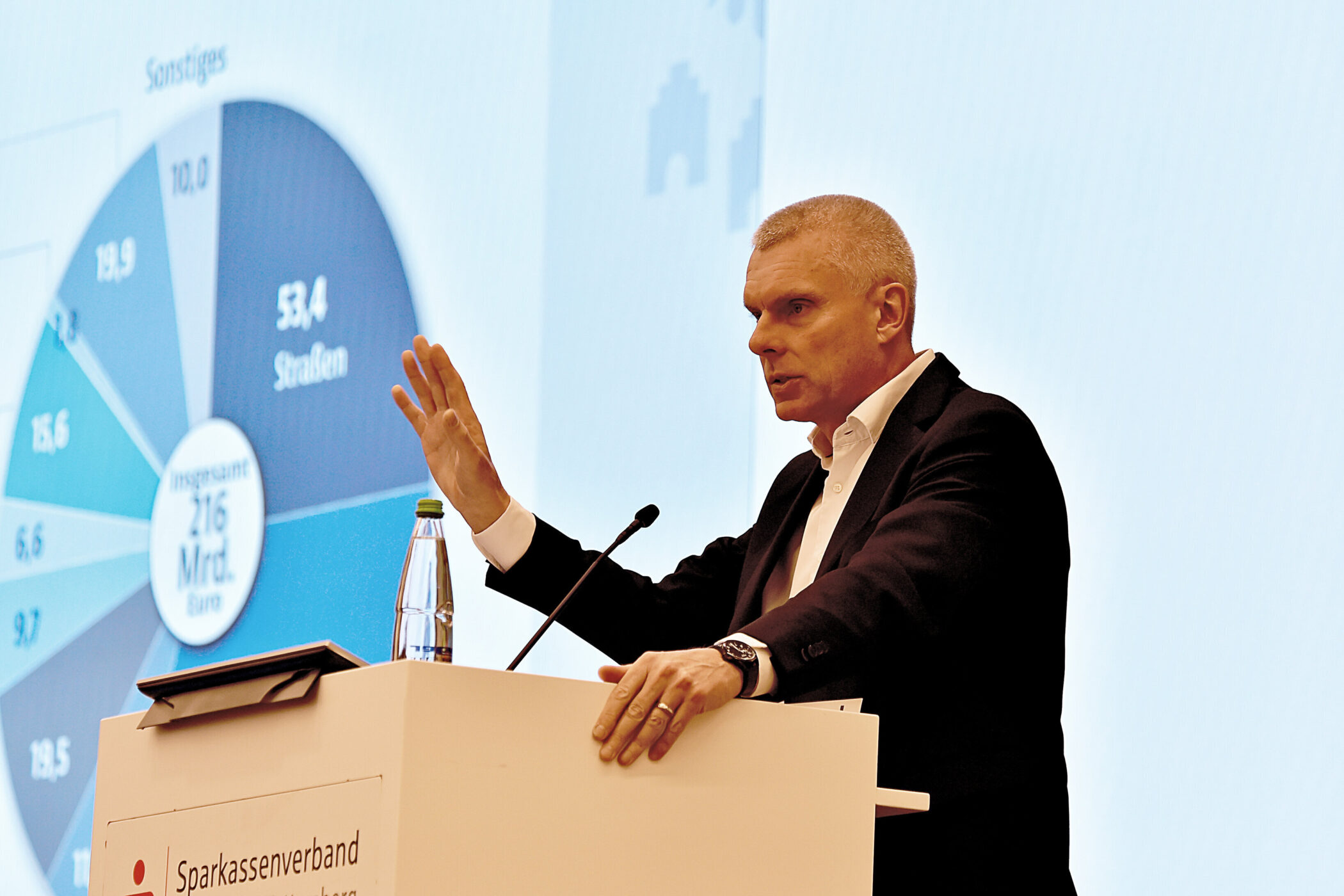 Mann am Rednerpult, zeigt auf Diagramm mit Finanzdaten.