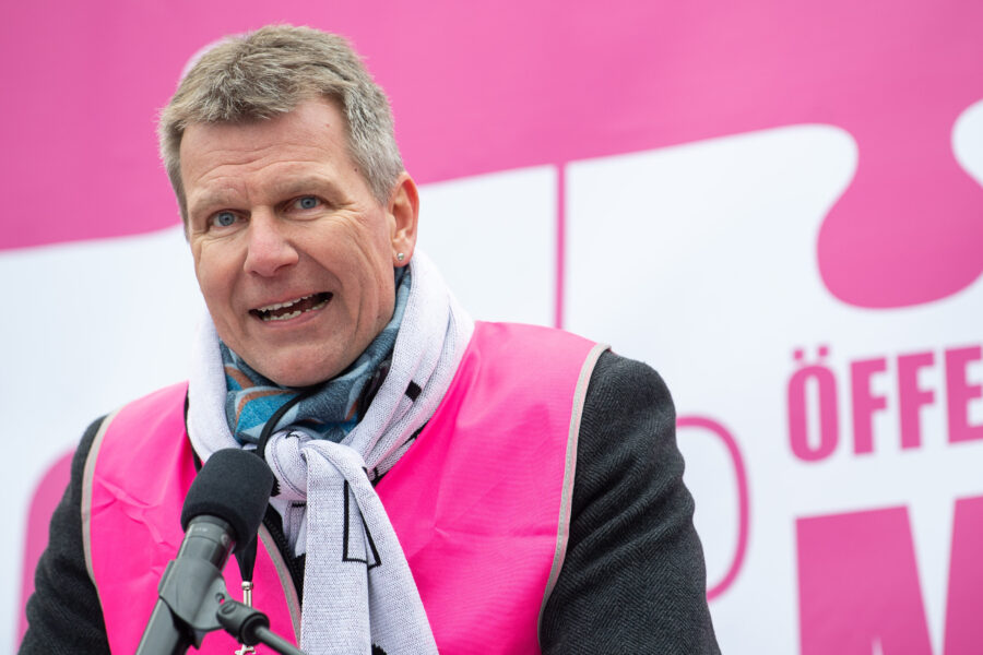 Mann mit Mikrofon, trägt pinke Weste und Schal, spricht vor pinkem Hintergrund.