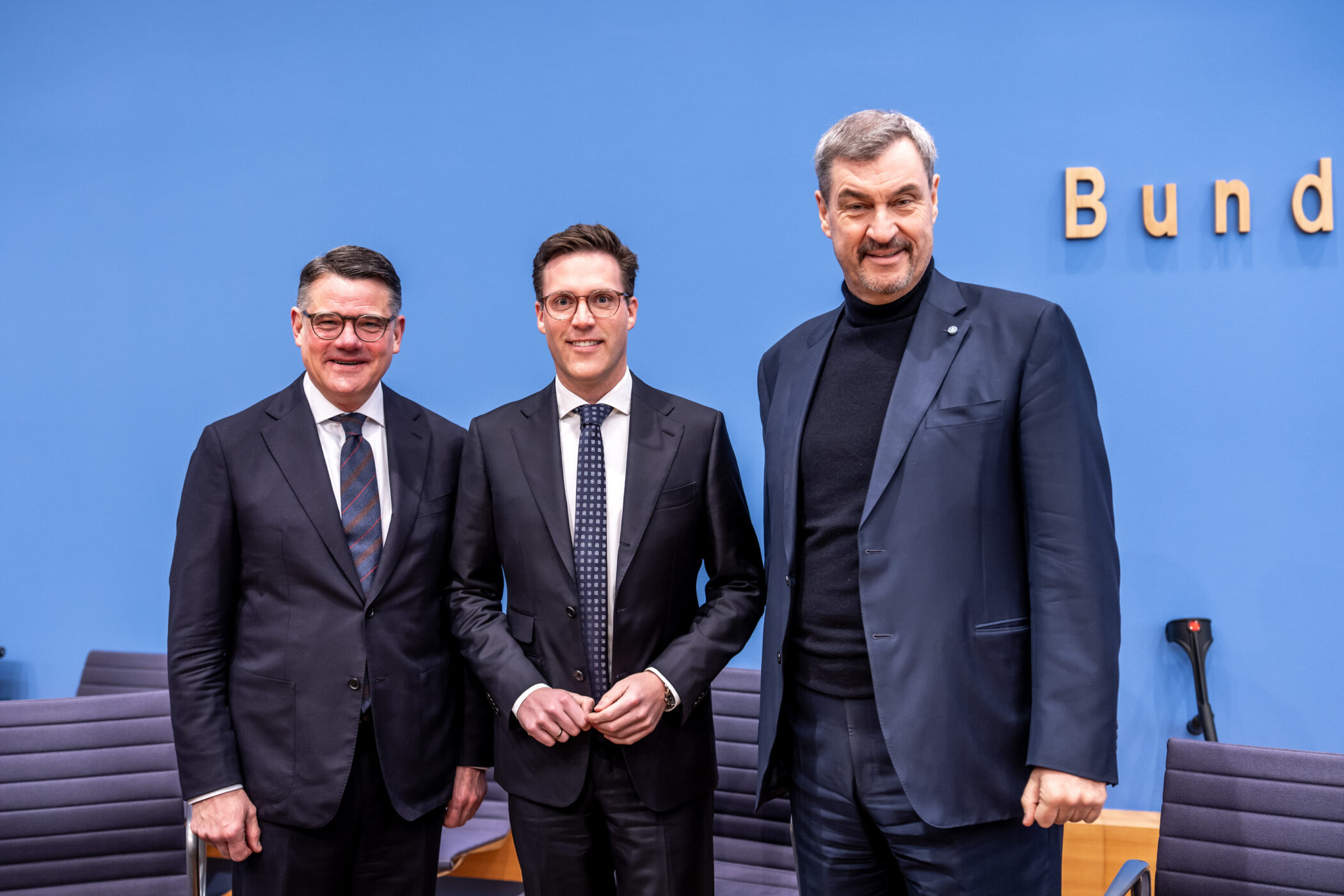 Drei Männer in Anzügen vor blauer Wand mit Schriftzug "Bund".