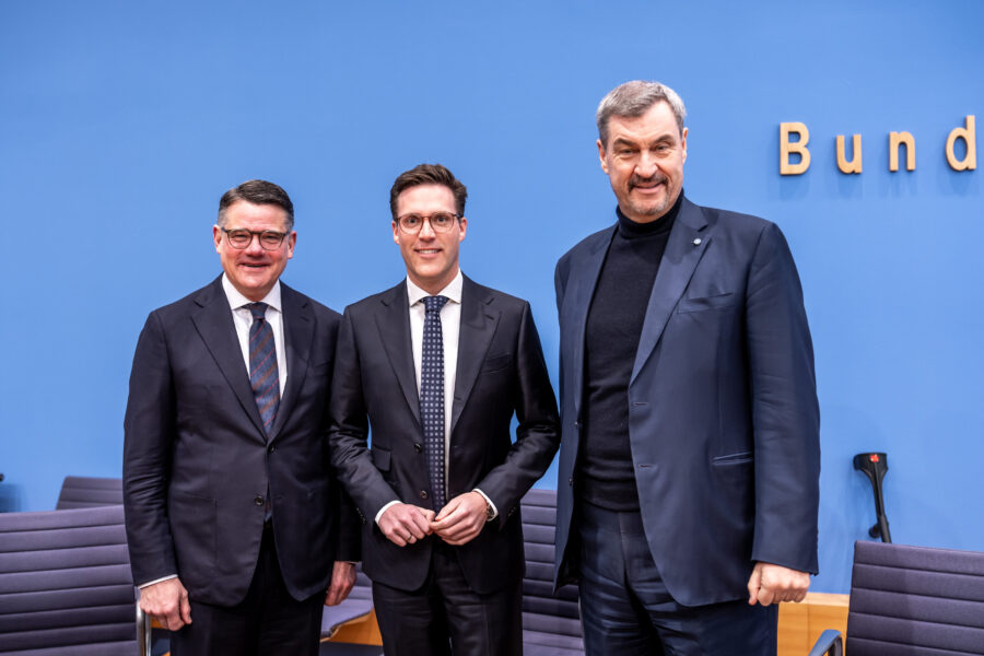 Drei Männer in Anzügen vor blauer Wand mit Schriftzug "Bund".