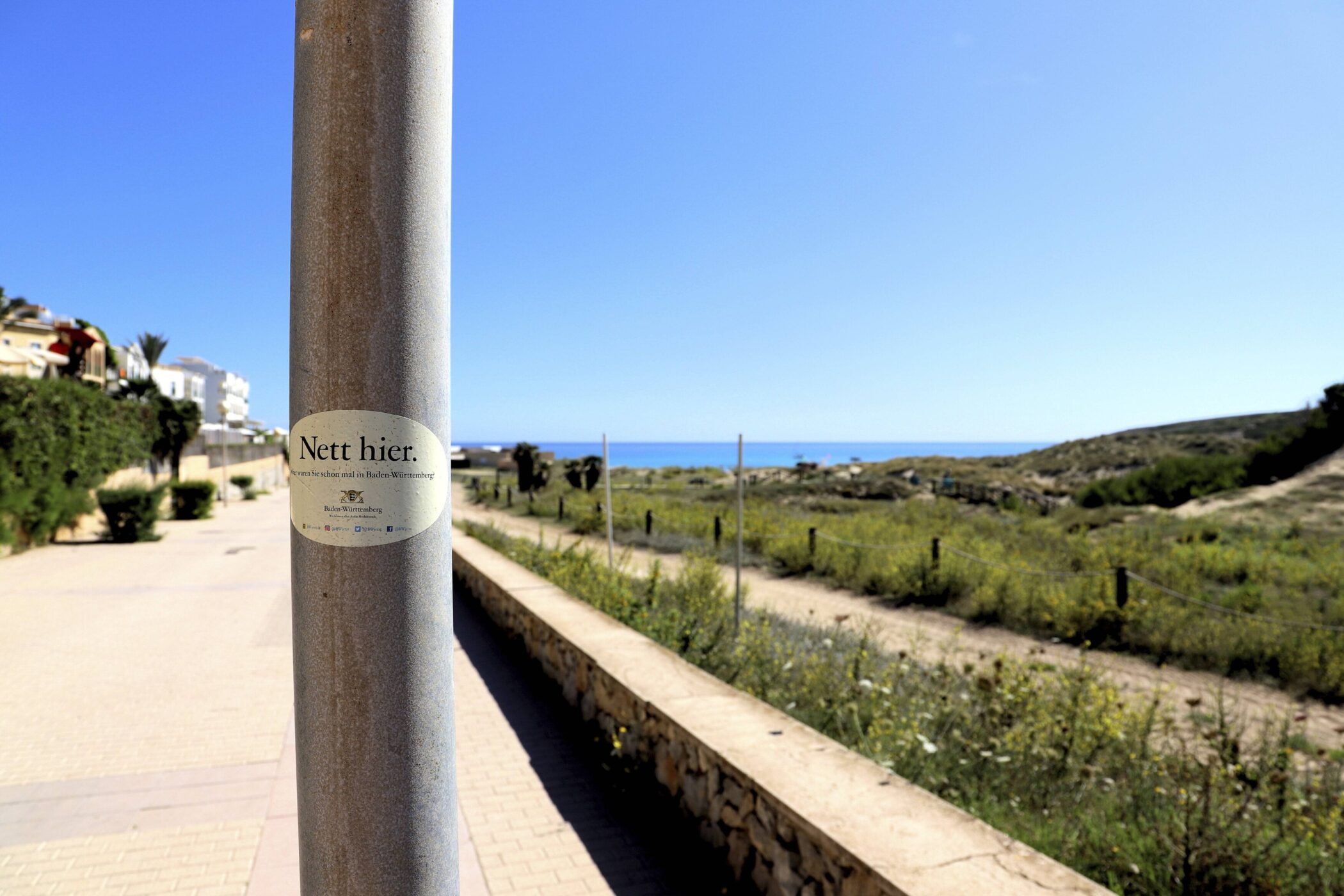 Laterne mit Aufkleber "Nett hier." vor Küstenlandschaft und blauem Himmel.