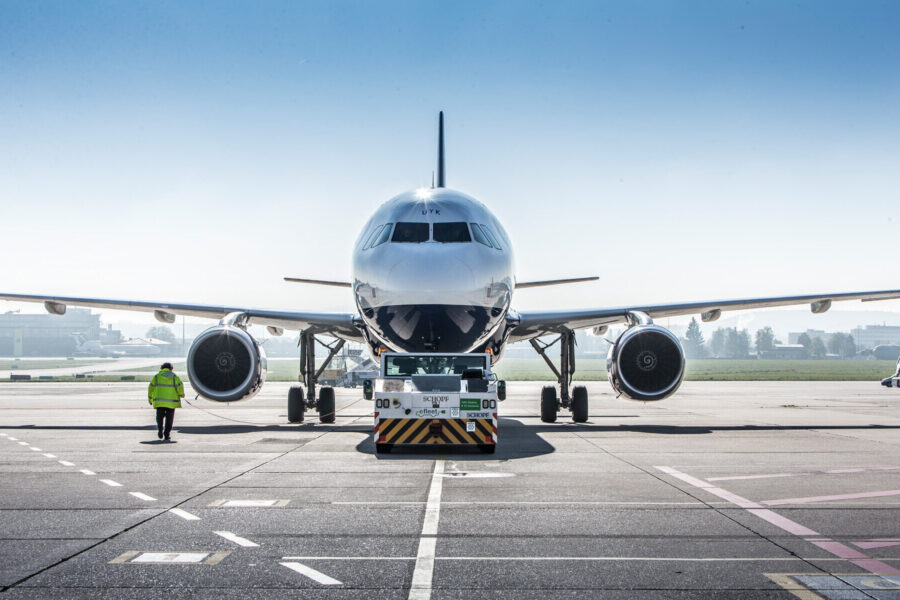 Flugzeug auf Rollfeld, Schlepper vorne, Person in gelber Weste daneben.