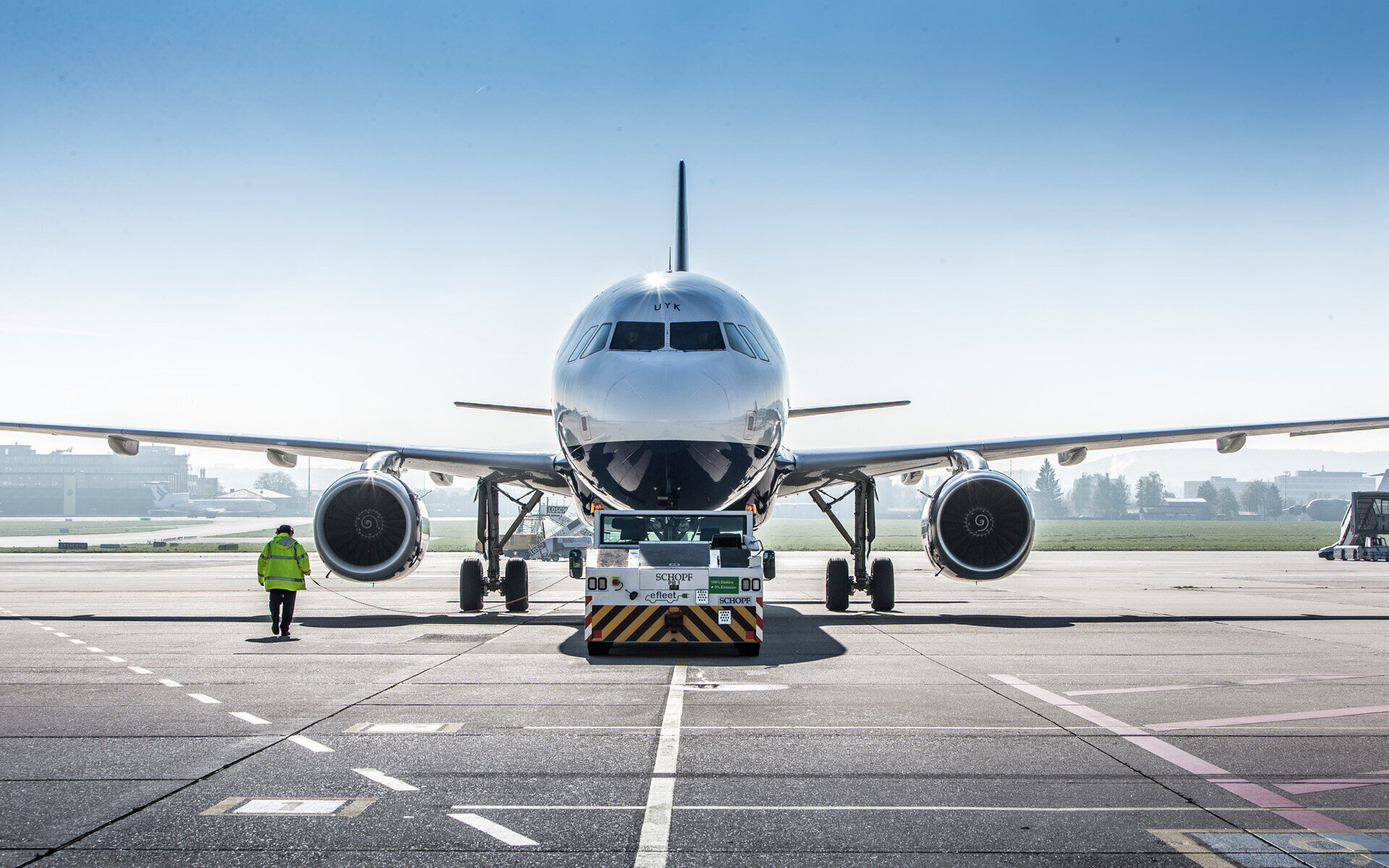 Flugzeug auf Rollfeld, Schlepper vorne, Person in gelber Weste daneben.