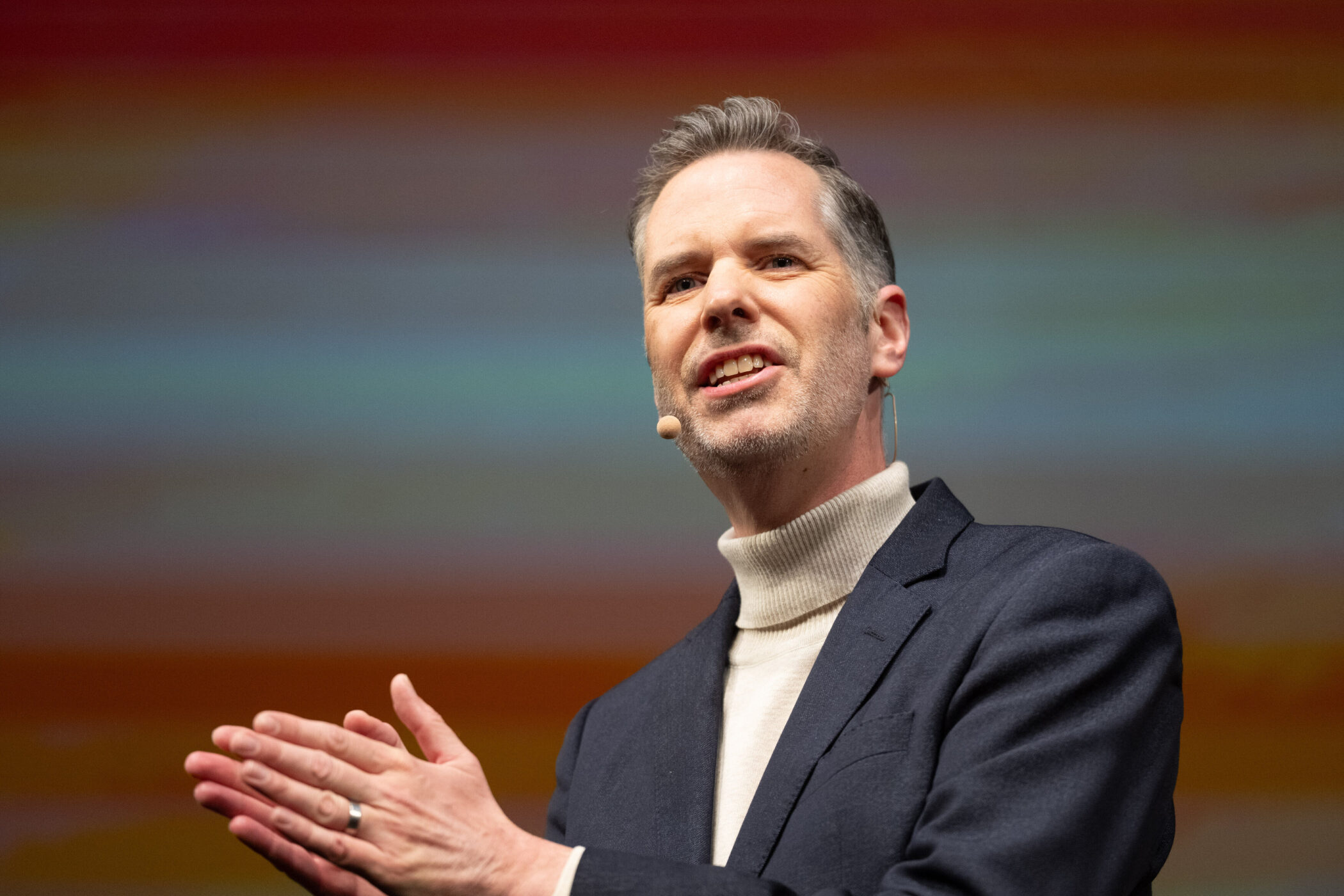 Mann mit Mikrofon spricht, trägt Anzug und Rollkragenpullover.