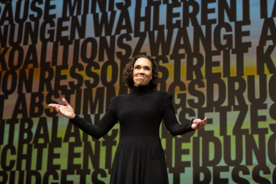 Person in schwarzem Kleid auf Bühne, Hintergrund mit großen Buchstaben.