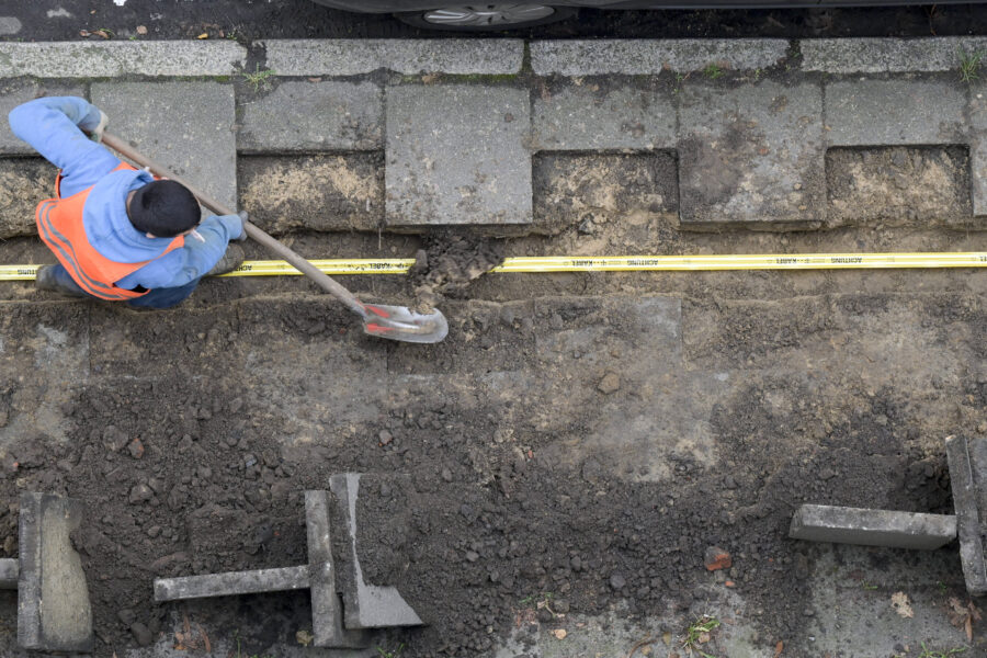 Person gräbt mit Schaufel einen Graben neben Gehwegplatten.