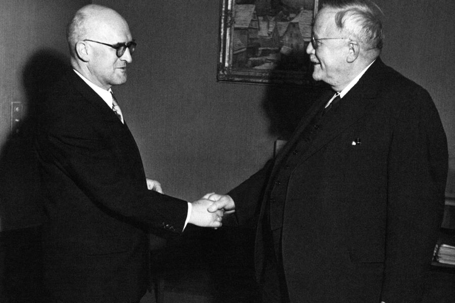 Zwei Männer im Anzug geben sich die Hand in einem Raum mit Gemälde an der Wand.