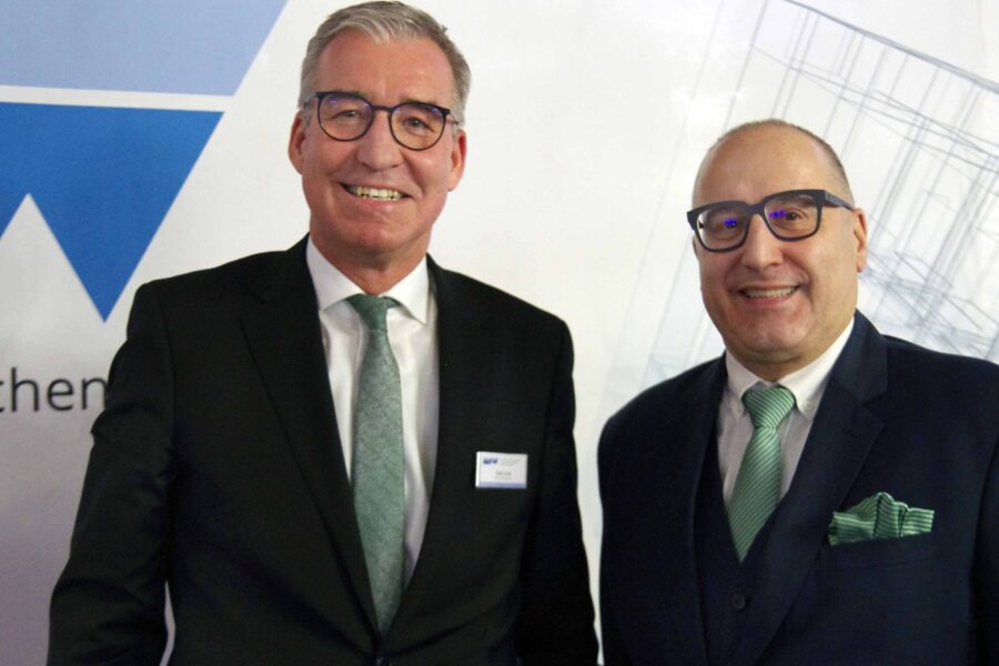 Zwei Männer in Anzügen lächeln in die Kamera, Hintergrund mit Logo.