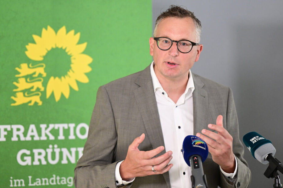 Mann im Anzug spricht vor Mikrofonen, grüner Hintergrund mit Sonnenblumen-Logo.