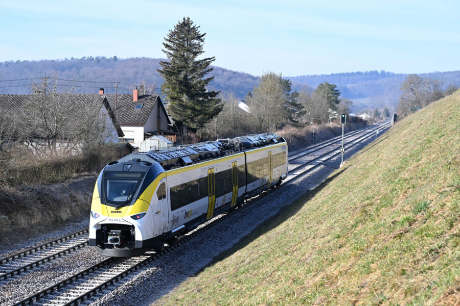 Zug auf zweigleisiger Strecke, Häuser und Bäume im Hintergrund, grünes Signal.