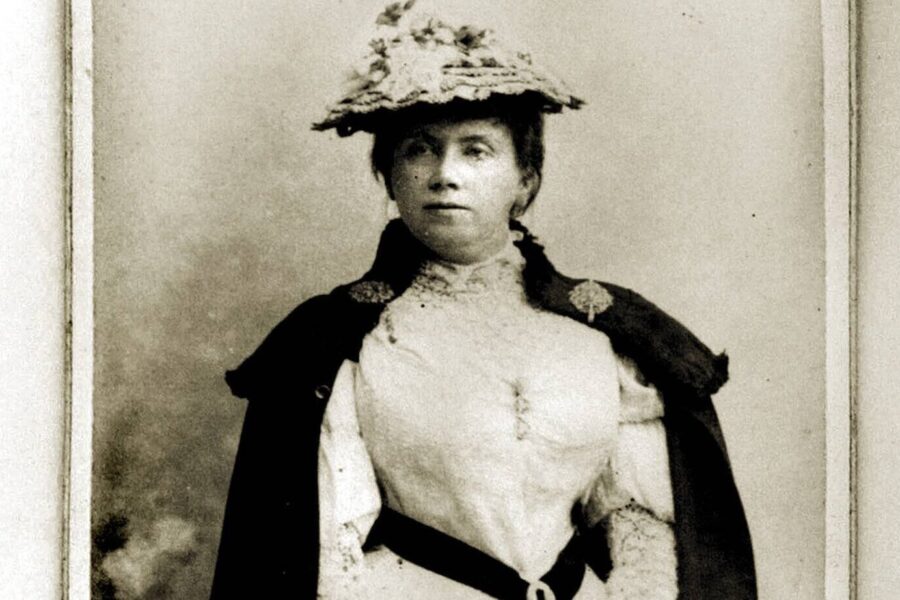 Frau in historischer Kleidung mit Hut und Umhang, frontal stehend.