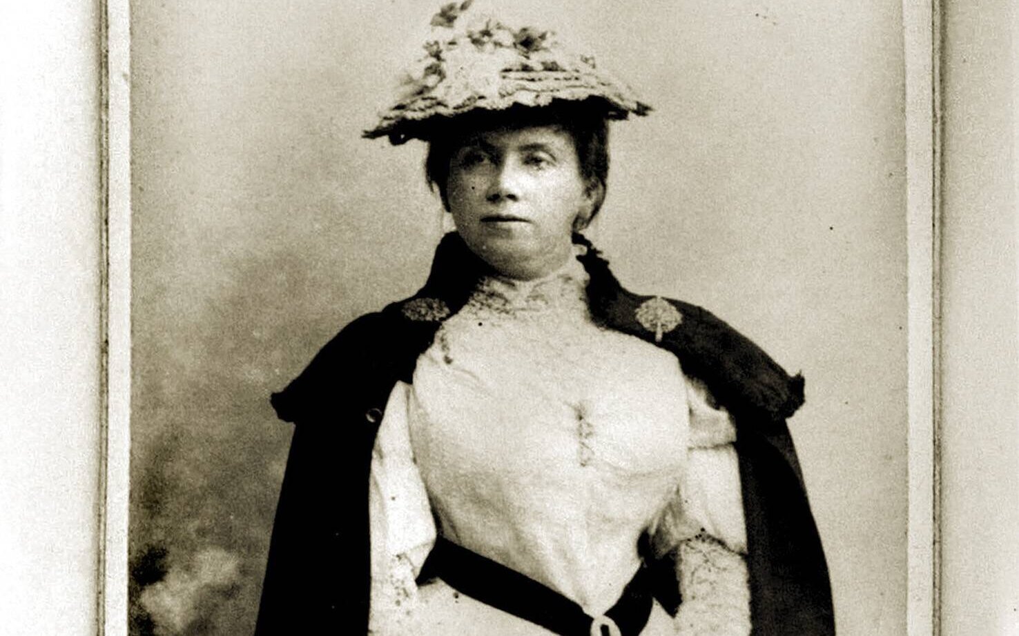 Frau in historischer Kleidung mit Hut und Umhang, frontal stehend.