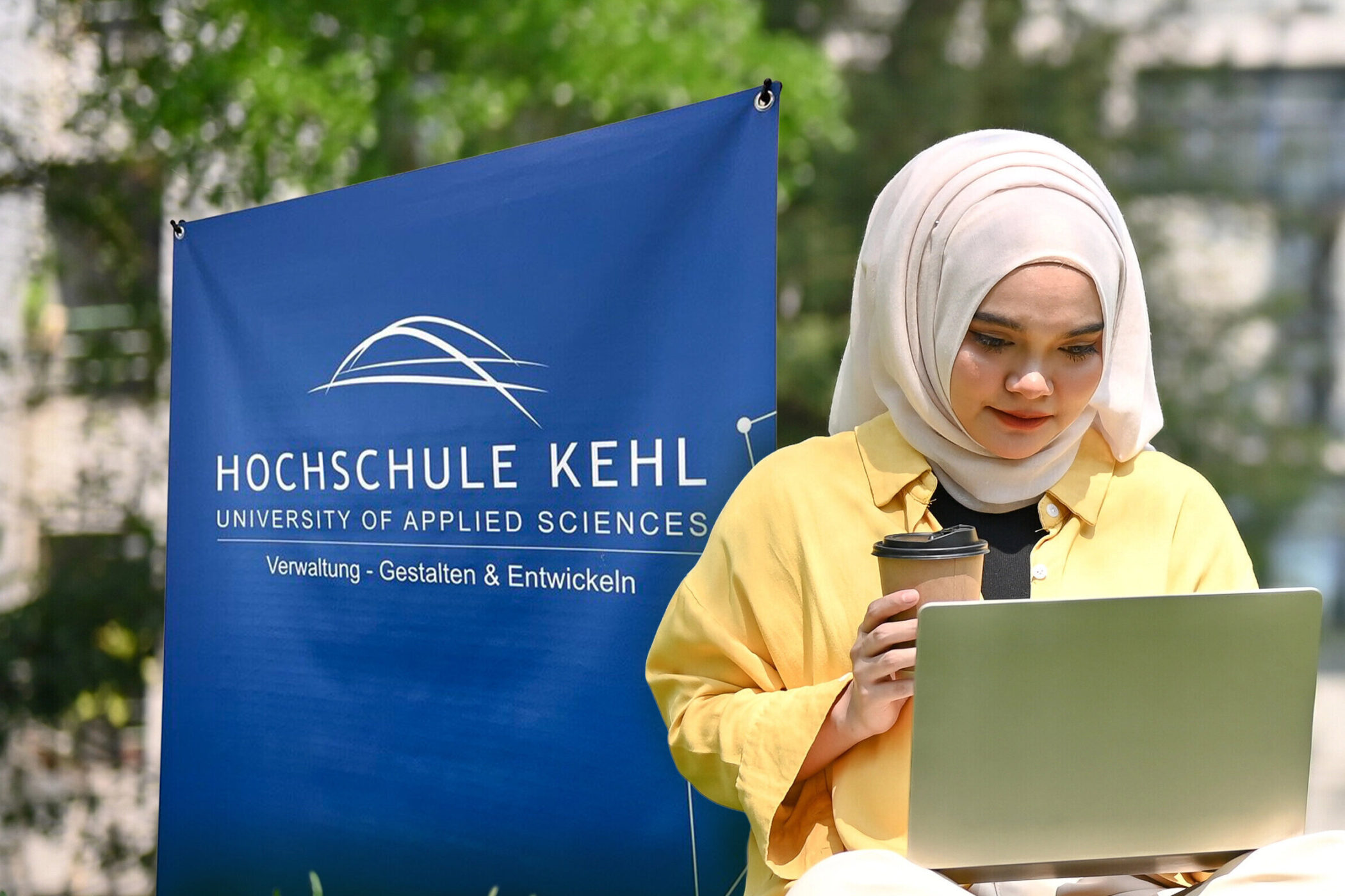 Frau mit Laptop und Kaffeebecher vor Banner der Hochschule Kehl im Freien.