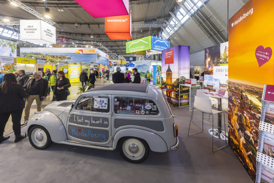 Messehalle mit Ständen, Menschen und einem grauen Auto mit "I lost my heart in Heidelberg".