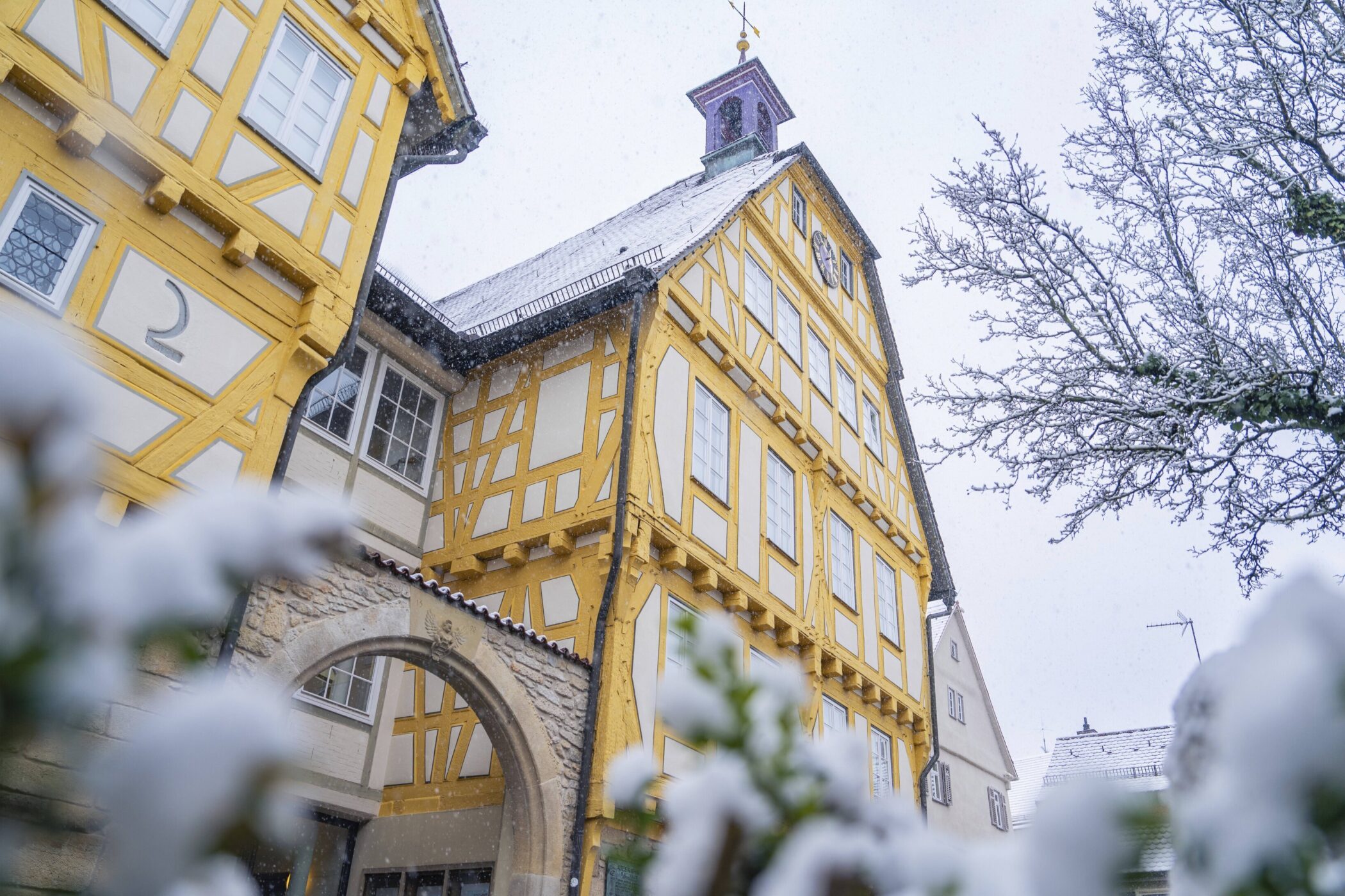Fachwerkhaus mit gelben Balken, Schnee auf Dach und Bäumen, bewölkter Himmel.