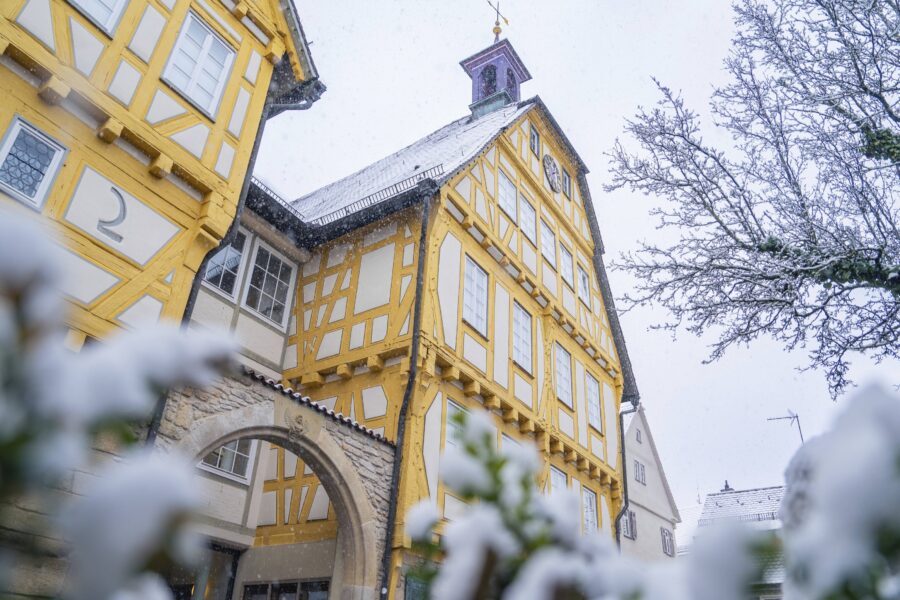 Fachwerkhaus mit gelben Balken, Schnee auf Dach und Bäumen, bewölkter Himmel.