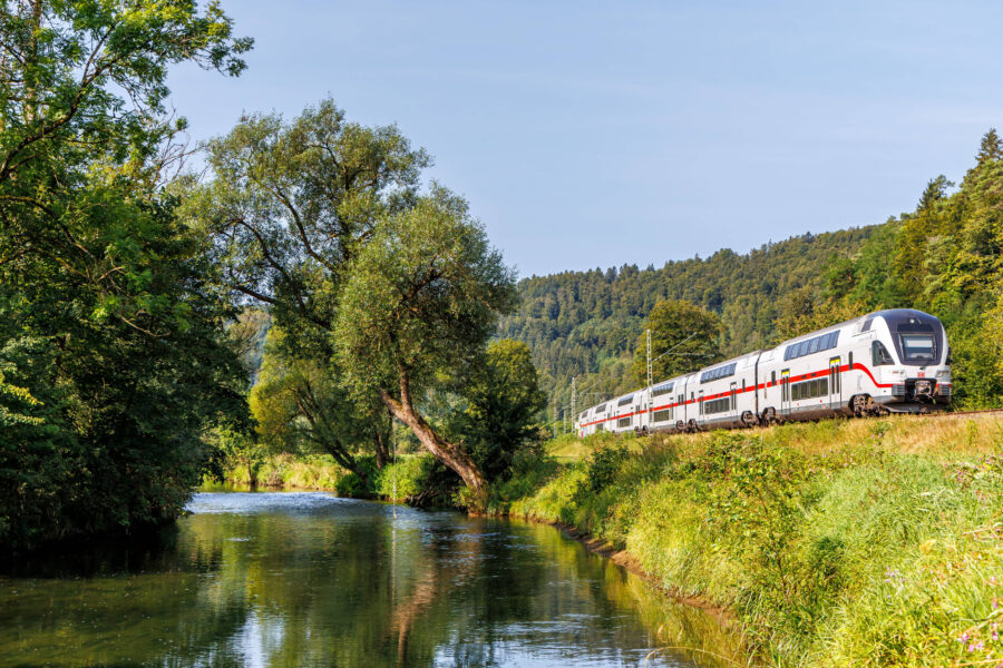 Zug fährt an einem Fluss entlang, umgeben von Bäumen und Hügeln.