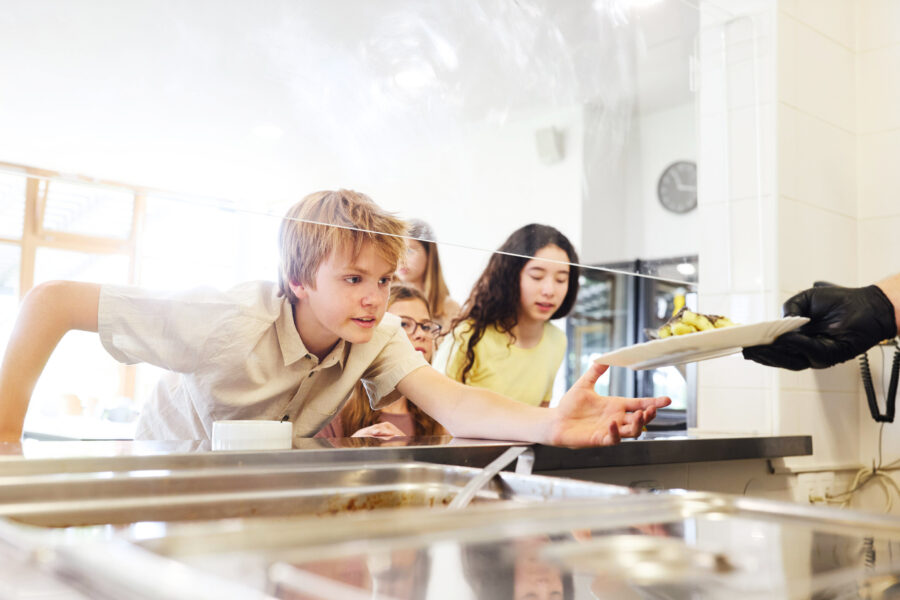 Vier Kinder an einer Essensausgabe, ein Kind nimmt einen Teller entgegen.