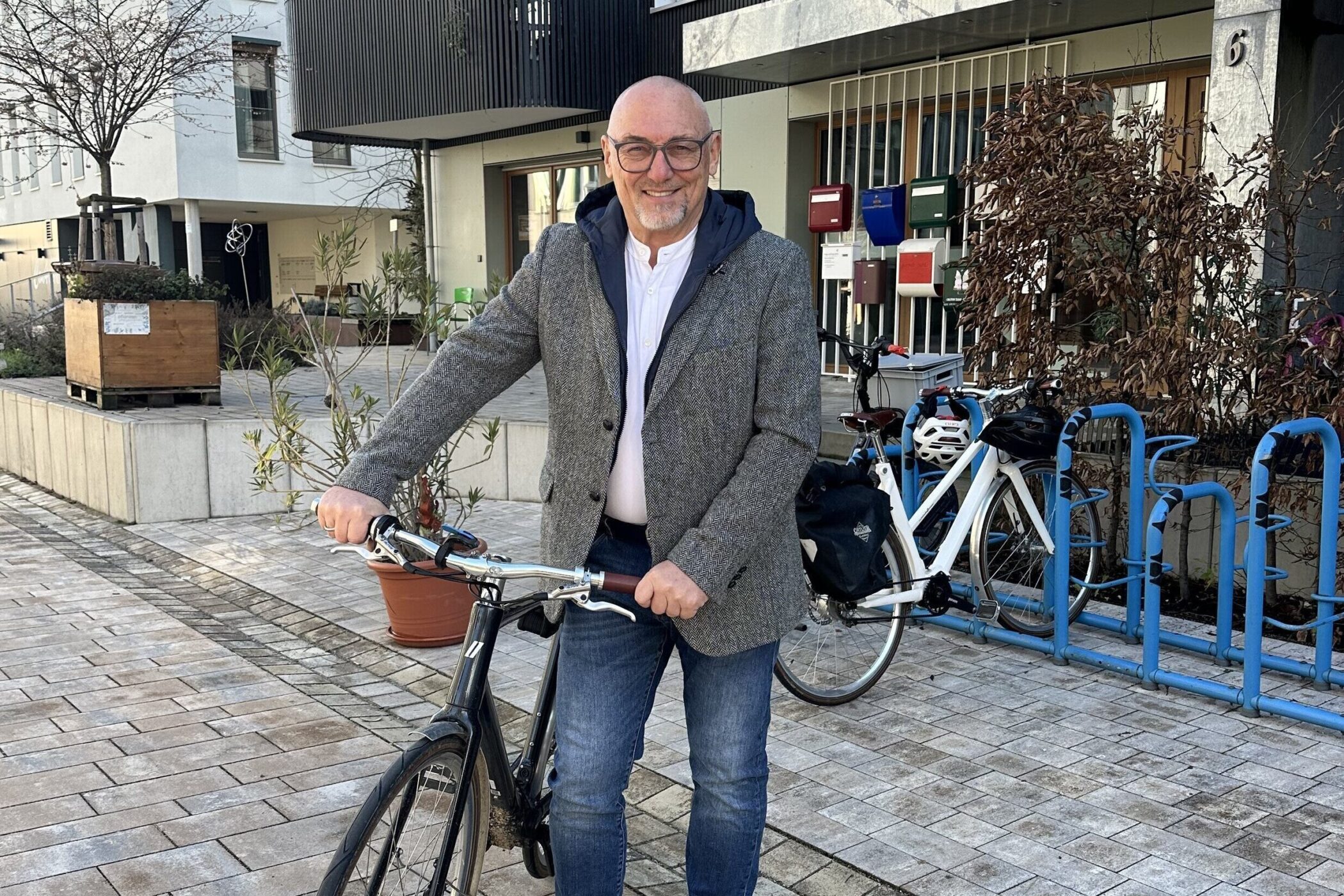 Mann mit Brille und grauem Sakko hält ein Fahrrad, im Hintergrund Fahrradständer.