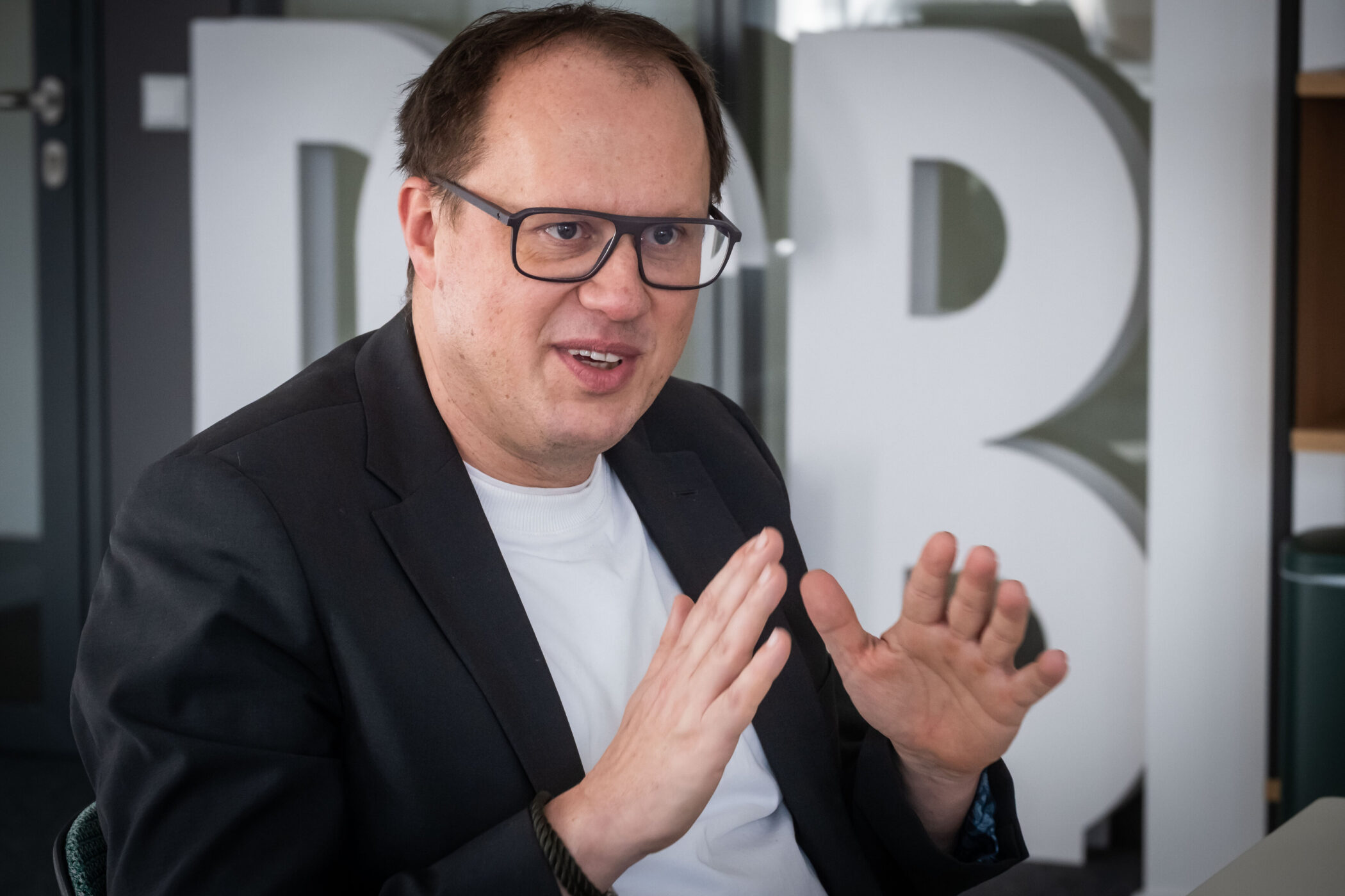 Ein Mann mit Brille gestikuliert, im Hintergrund große Buchstaben.